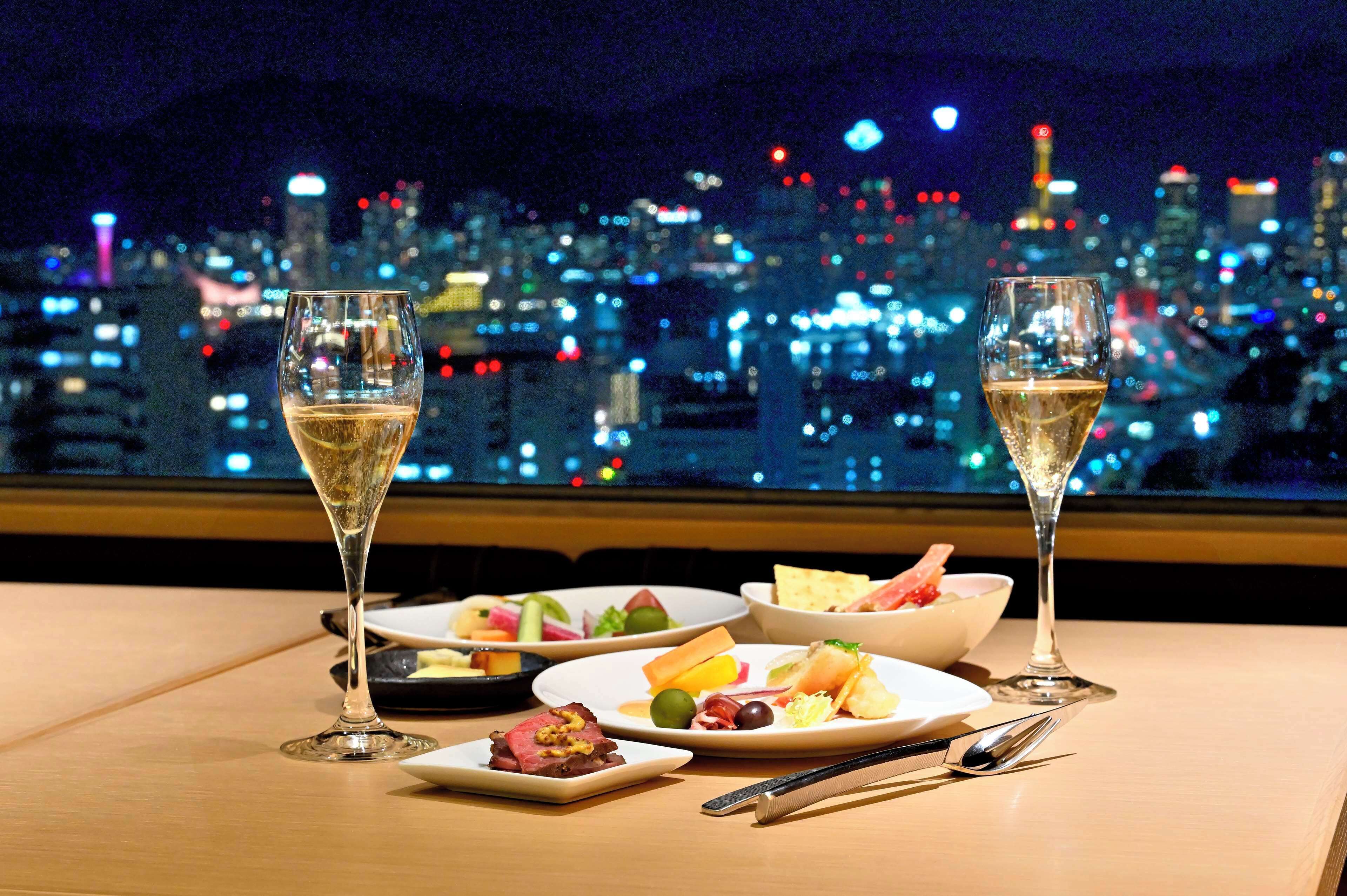 Une table avec deux coupes de champagne, divers plats et couverts, le tout mis en valeur par une vue sur la ville illuminée la nuit.