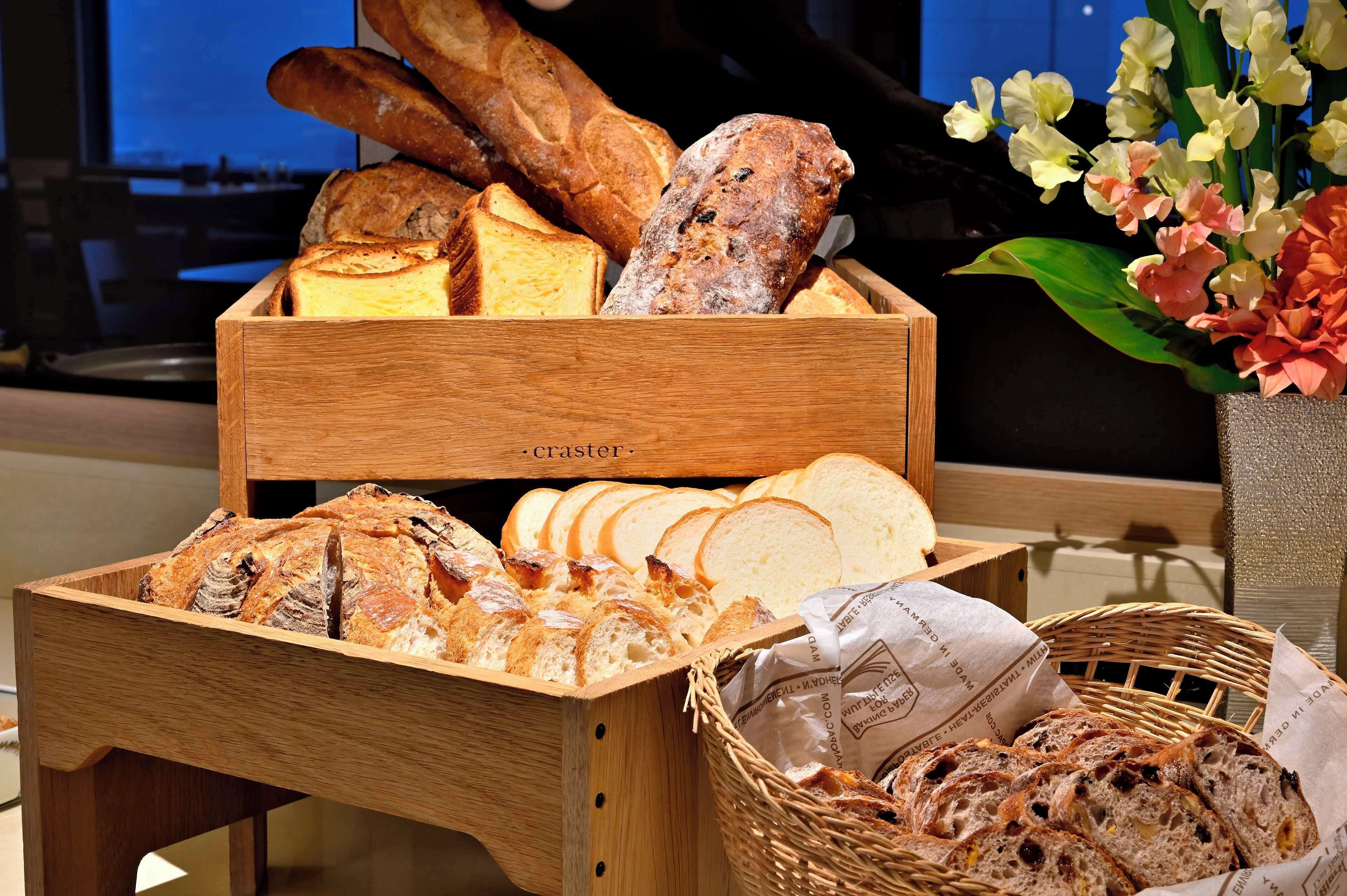 A wooden bread basket filled with various types of bread, a wicker basket with bread, and a vase with flowers.