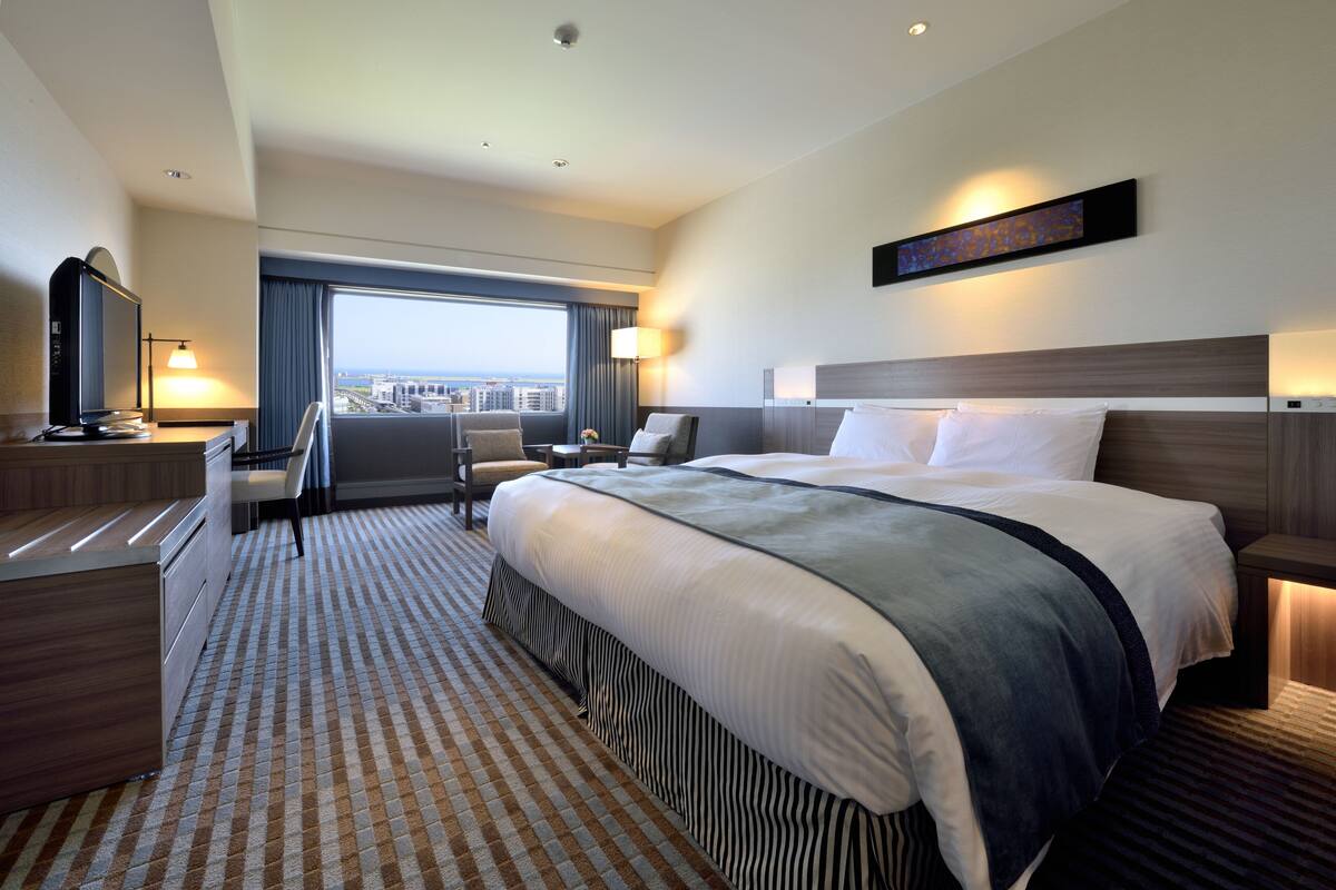 A hotel room with a large bed, a desk with a chair, a TV, and a view of the city.