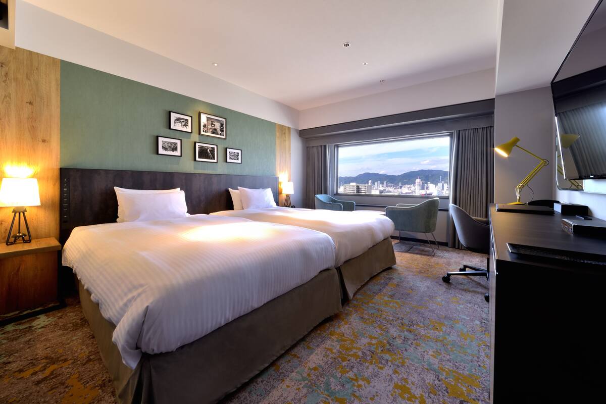 A hotel room with two beds, a desk, a chair, a view of the city, and framed pictures on the wall.