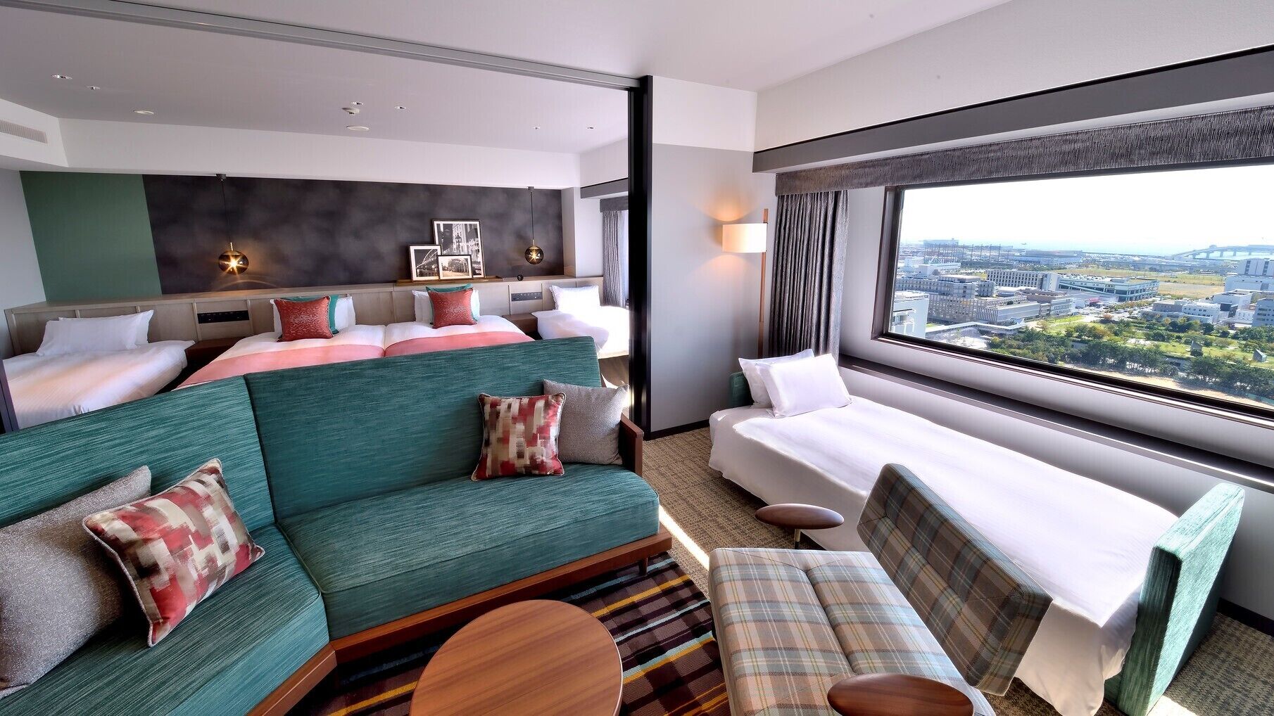 A hotel room with a bed, a sofa, a chair, a small table, and a large window with a city view.