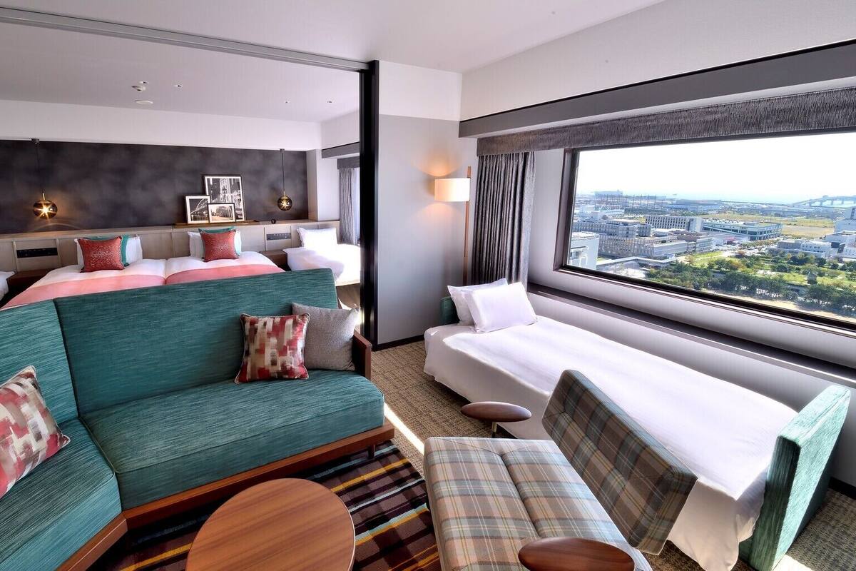 A hotel room with a bed, a sofa, a chair, a small table, and a large window with a city view.
