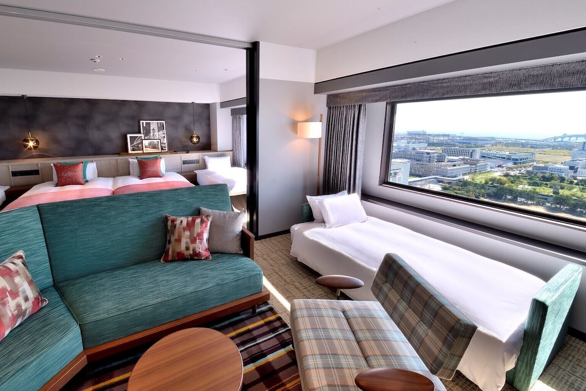 Ein Hotelzimmer mit einem Bett, einem Sofa, einem Sessel, einem kleinen Tisch und einem großen Fenster mit Blick auf die Stadt.