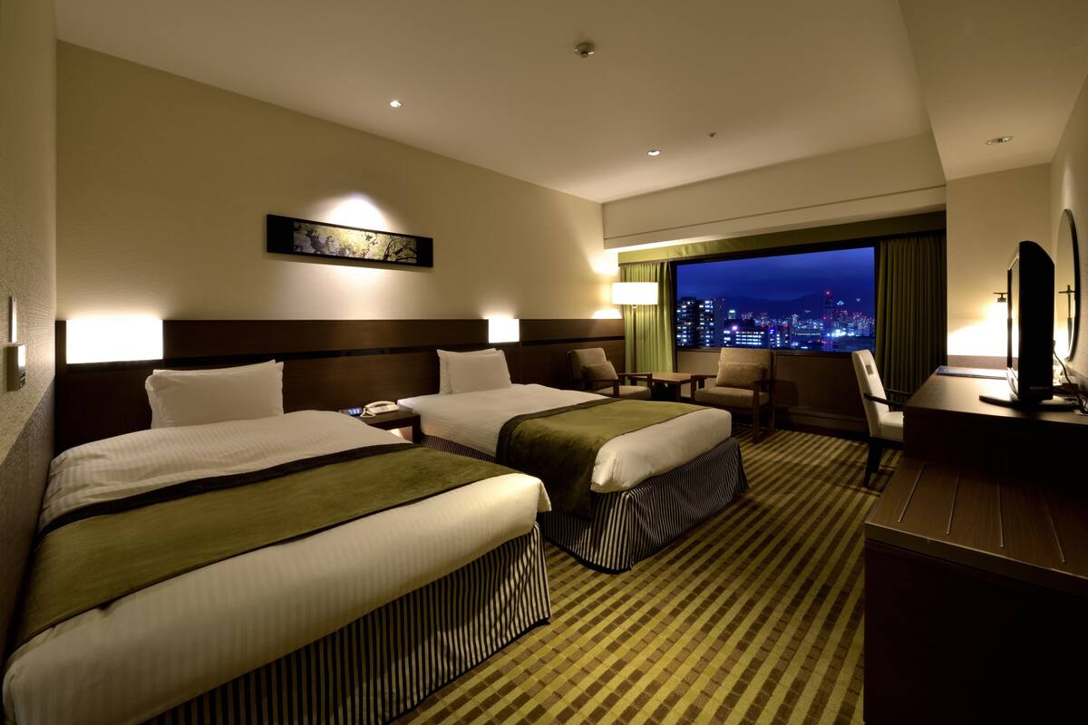 A hotel room with two beds, a desk, a chair, a TV, and a city view.