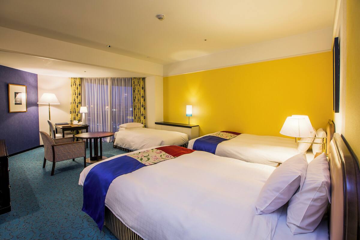 A hotel room with two beds, a desk, a chair, and a window with curtains.