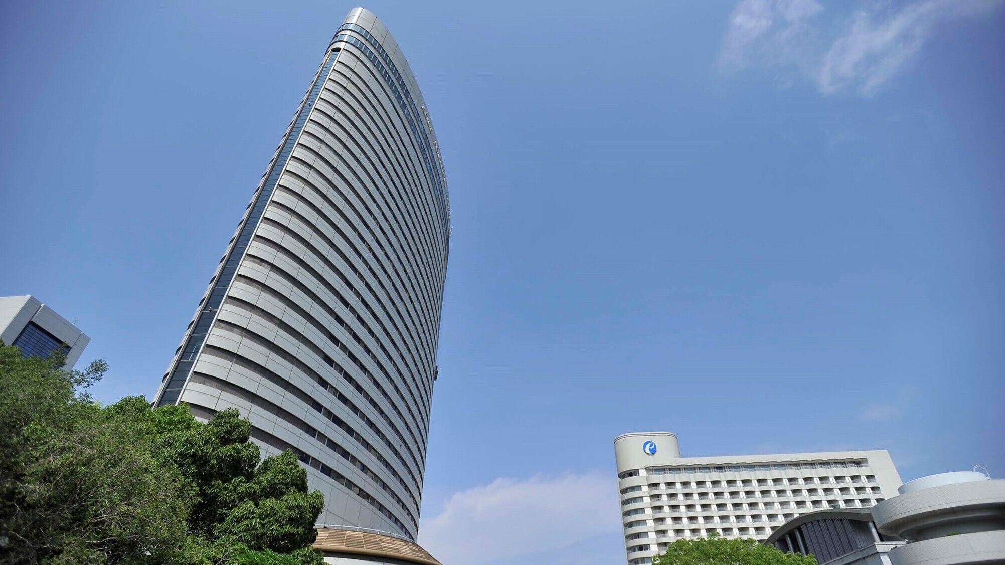 A tall, curved skyscraper with a modern design, surrounded by other buildings and trees.