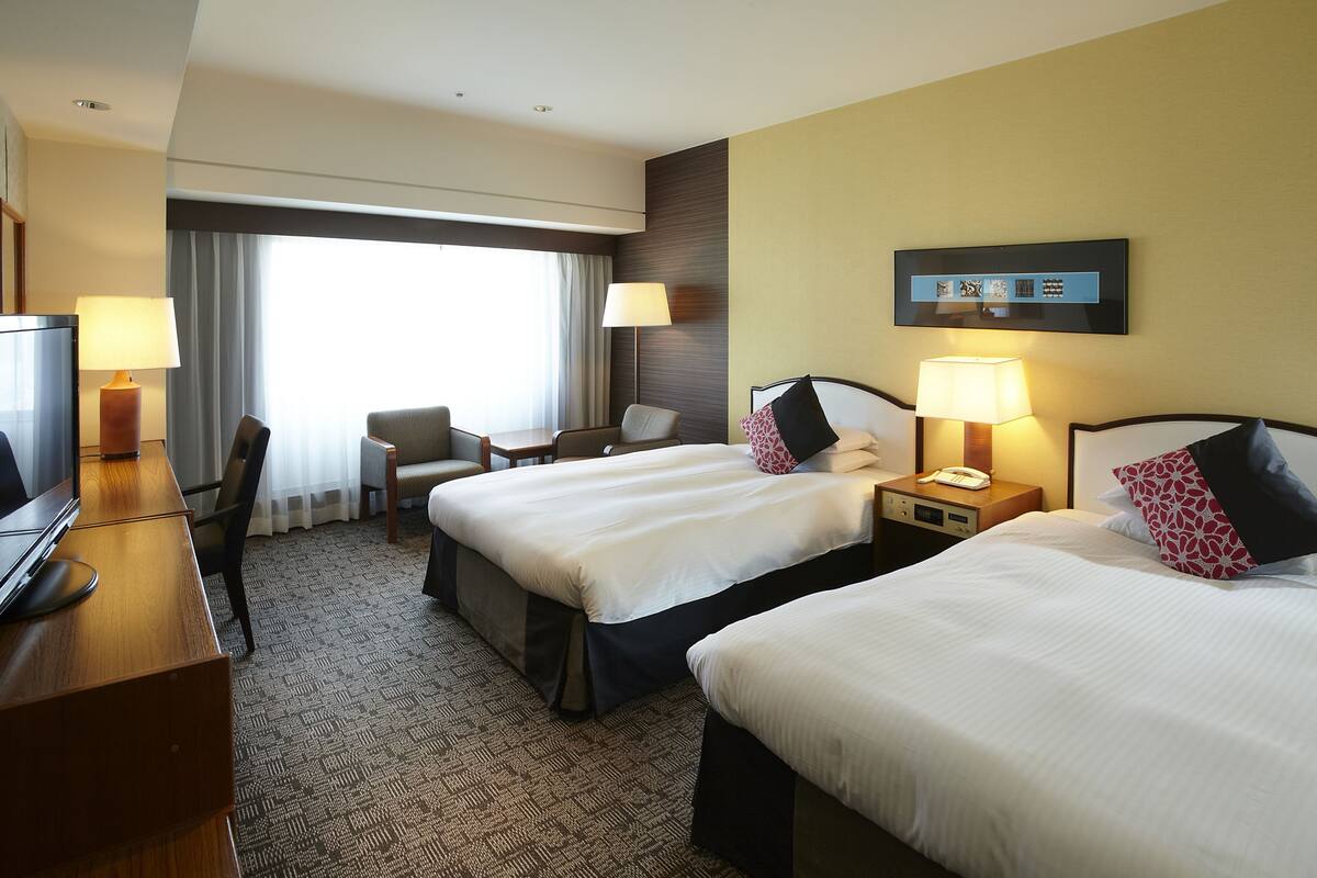 A hotel room with two beds, a desk with a TV, a chair, a lamp, and a window with curtains.