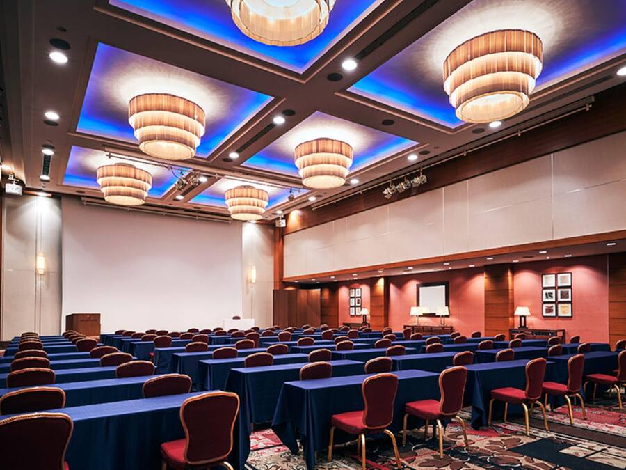 Un salón de conferencias con filas de mesas y sillas, un escenario y una pantalla grande.