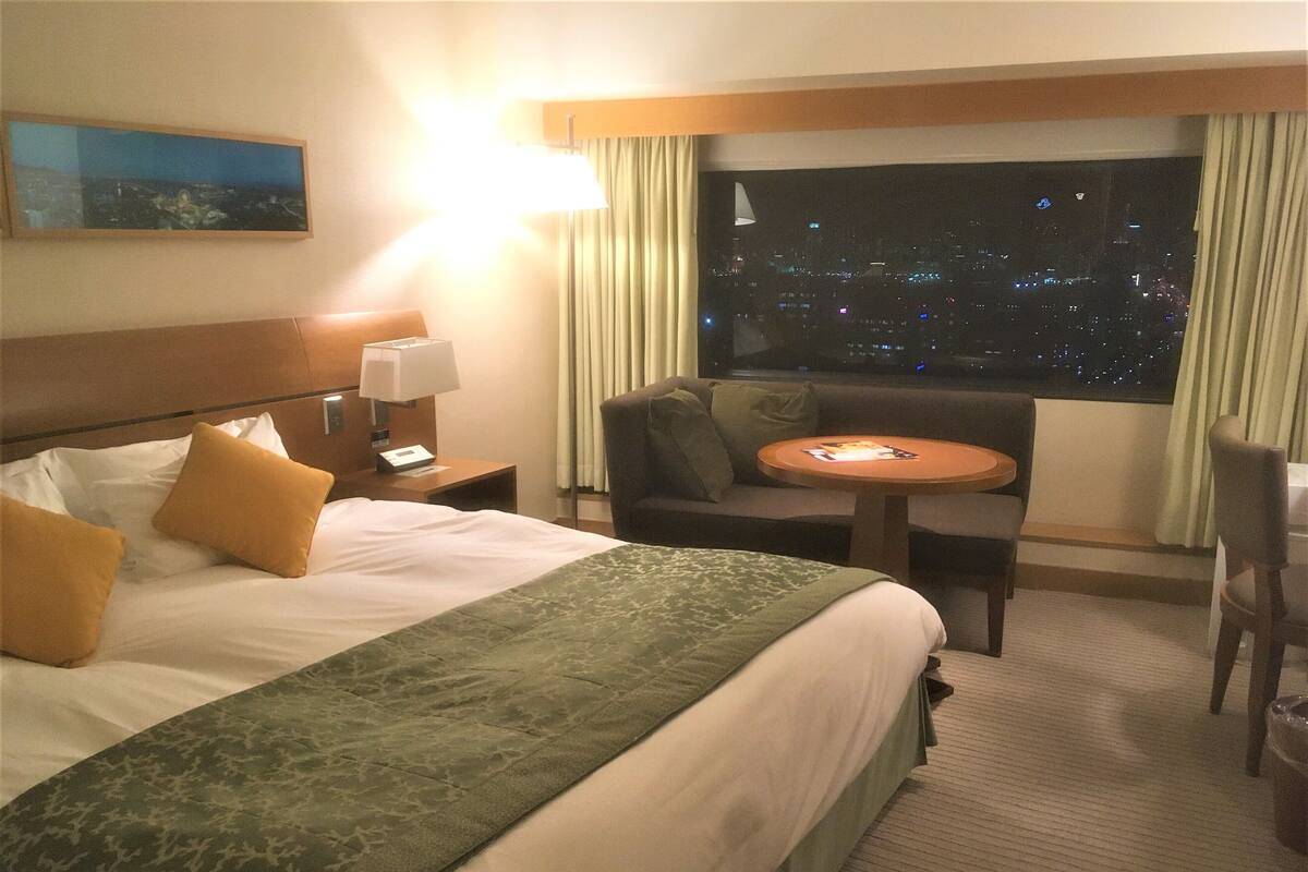 A hotel room with a bed, a nightstand, a lamp, a chair, a small table, and a view of a cityscape at night.