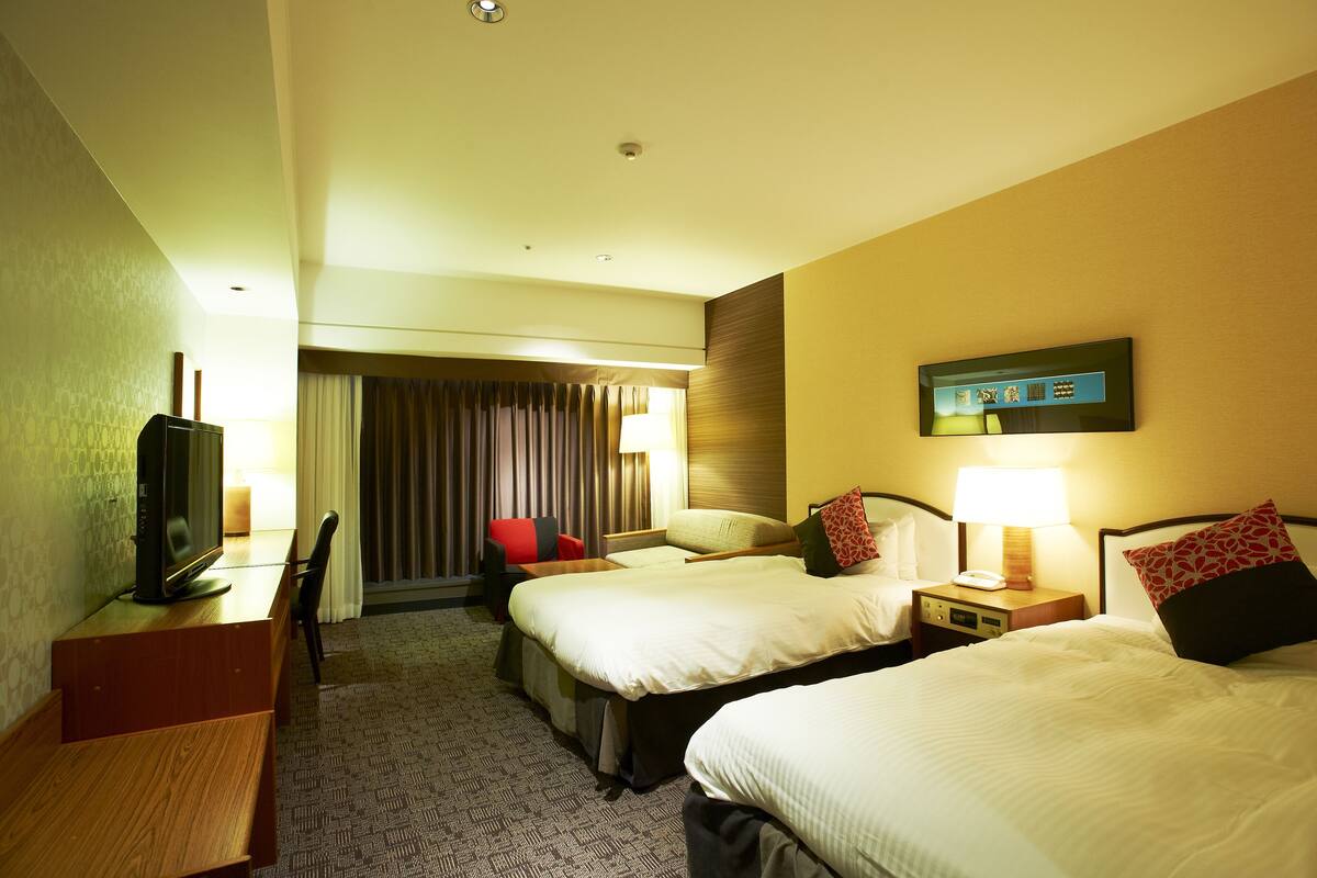 A hotel room with two beds, a desk with a television, a chair, and a lamp.