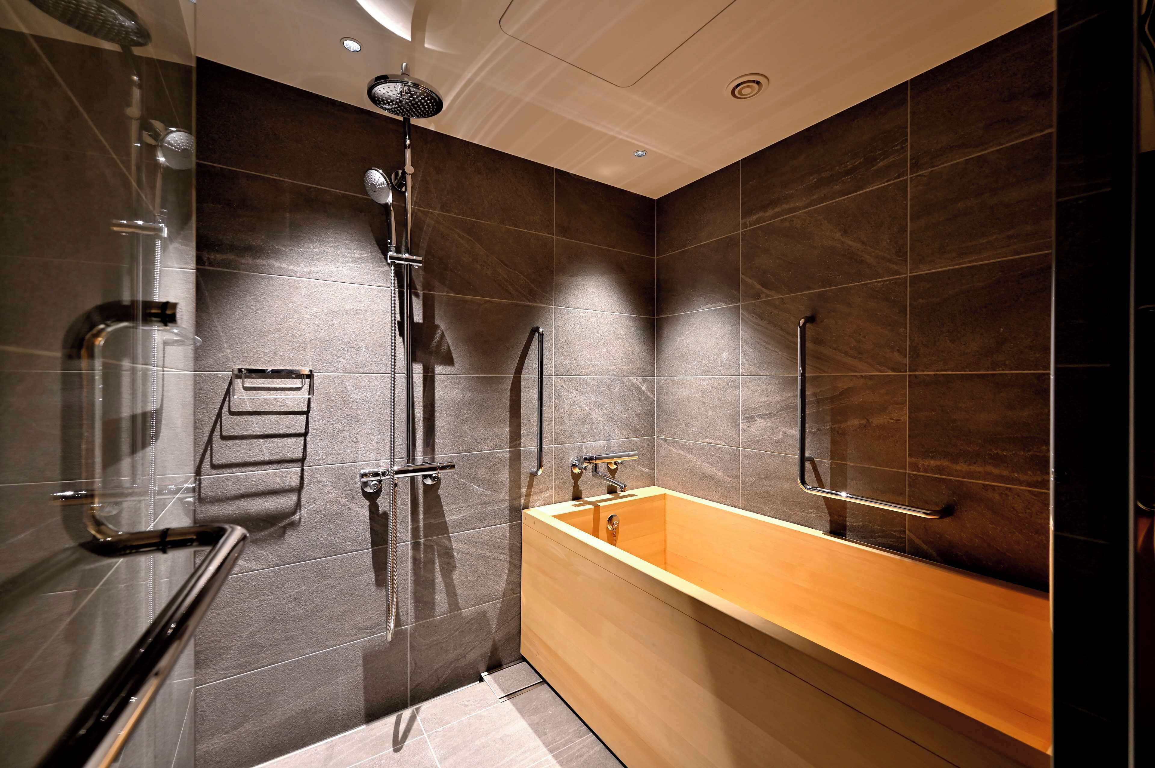 A modern bathroom with a glass shower enclosure, a wooden bathtub, and dark tiled walls.