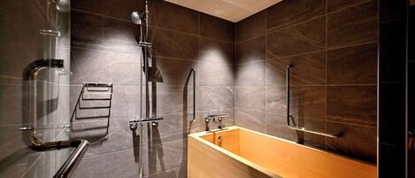 A modern bathroom with a glass shower enclosure, a wooden bathtub, and dark tiled walls.