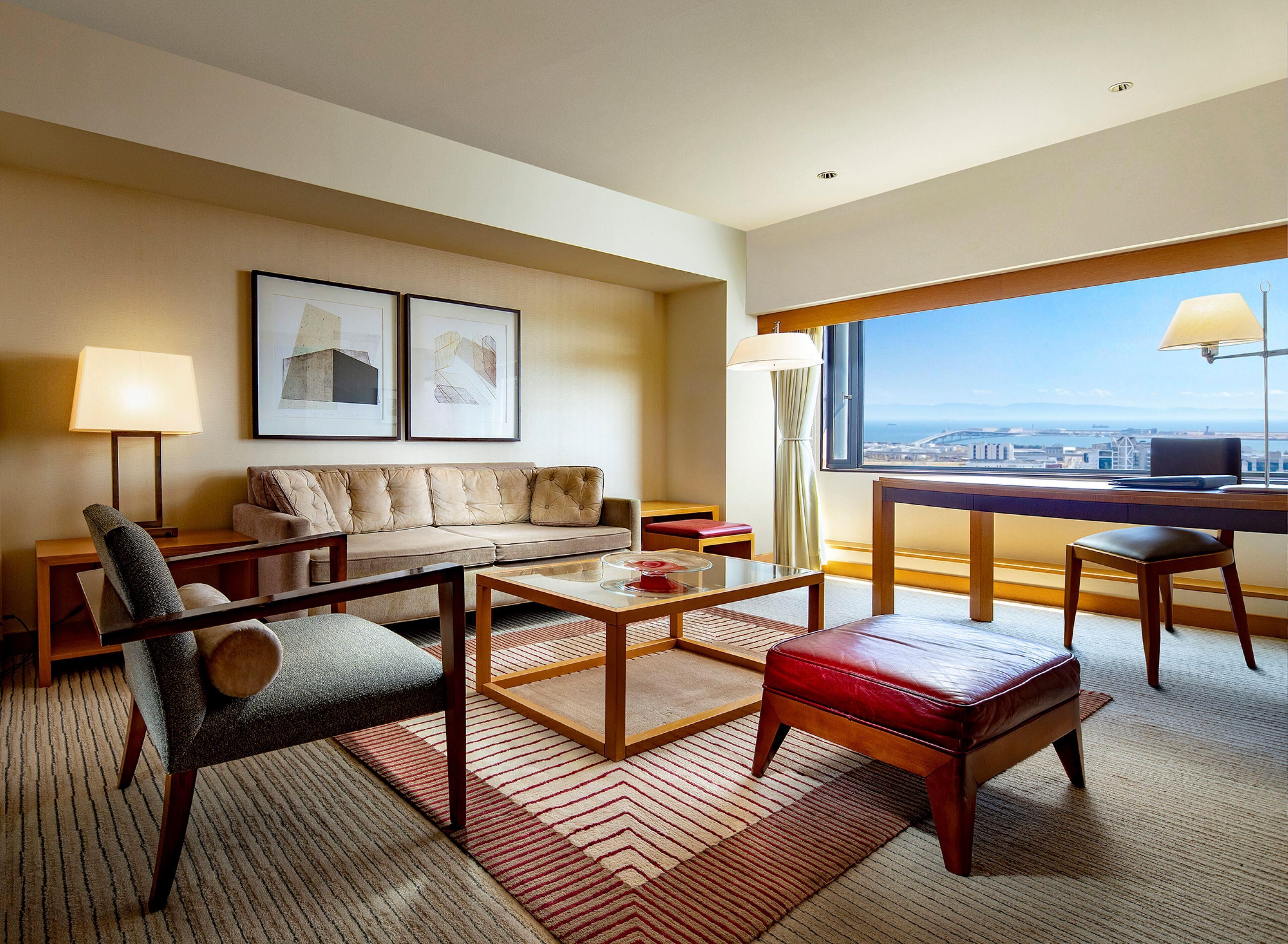 Habitación de hotel con sofá, sillón, mesa de centro y escritorio con vistas a la ciudad.