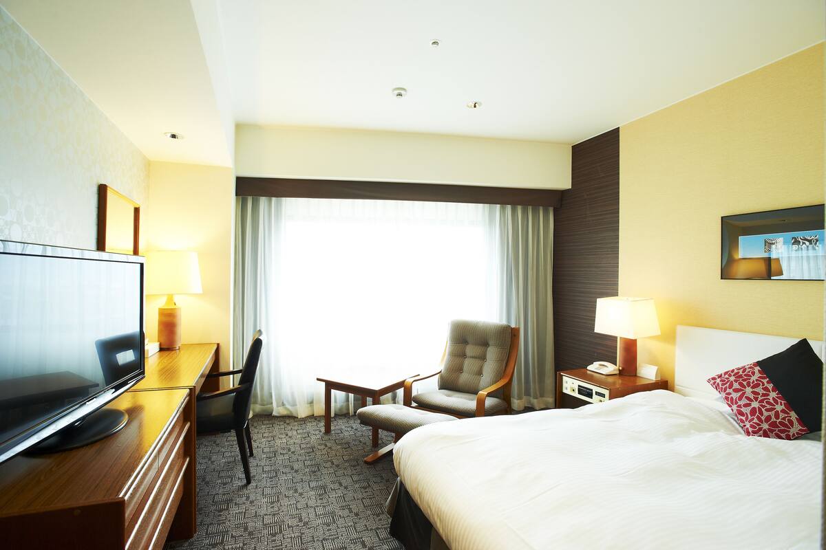 A hotel room with a bed, a desk with a television, a chair, a nightstand with a lamp, and a window with curtains.