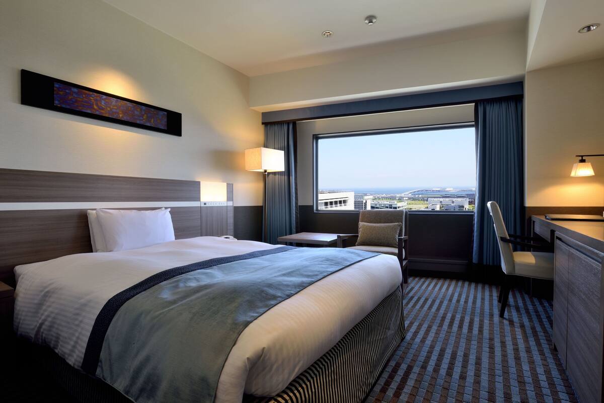 A hotel room with a large bed, a desk, a chair, a view of the city, and a painting on the wall.