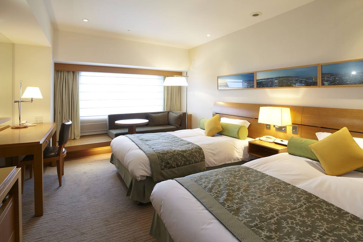 A hotel room with two beds, a desk, a chair, a nightstand, and a window with curtains.