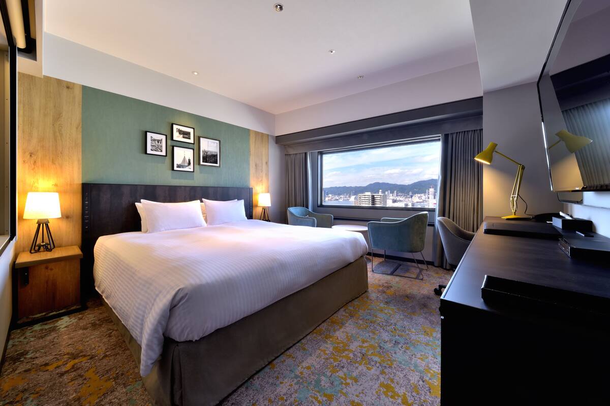 A hotel room with a large bed, a desk, a chair, a lamp, and a view of the city through a large window.