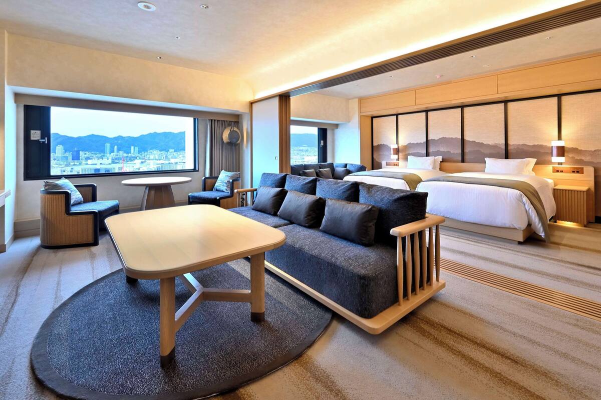 A modern hotel room with a large bed, a sofa, a dining table, and a view of the cityscape.