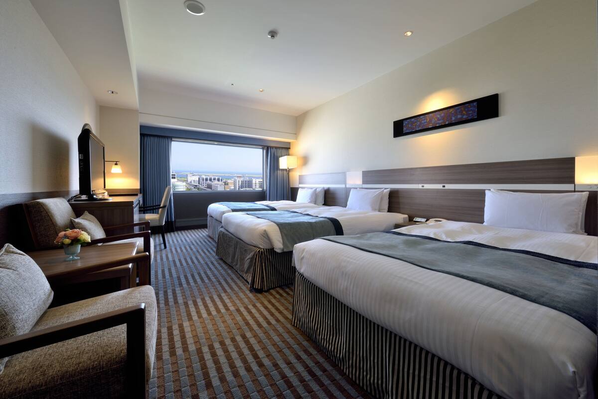 A hotel room with two beds, a desk with a lamp, a chair, and a view of the city.