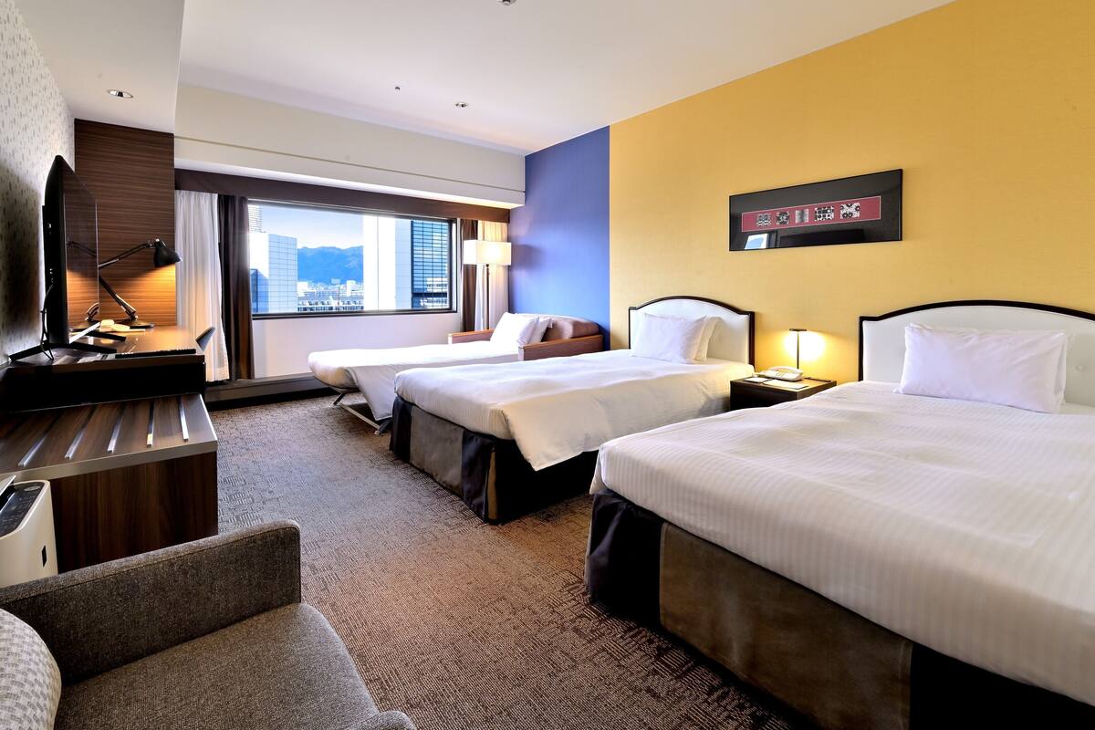 A hotel room with two beds, a desk, a chair, a TV, and a window with a city view.
