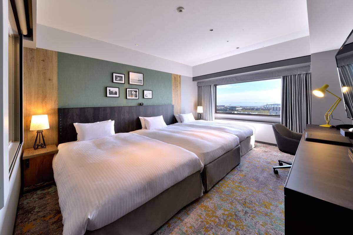 A hotel room with two beds, a desk, a chair, a large window with a view, and a lamp.