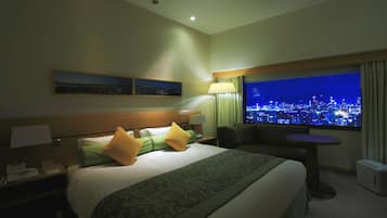 A hotel room with a bed, a nightstand, a lamp, a table, and a large window with a city view.