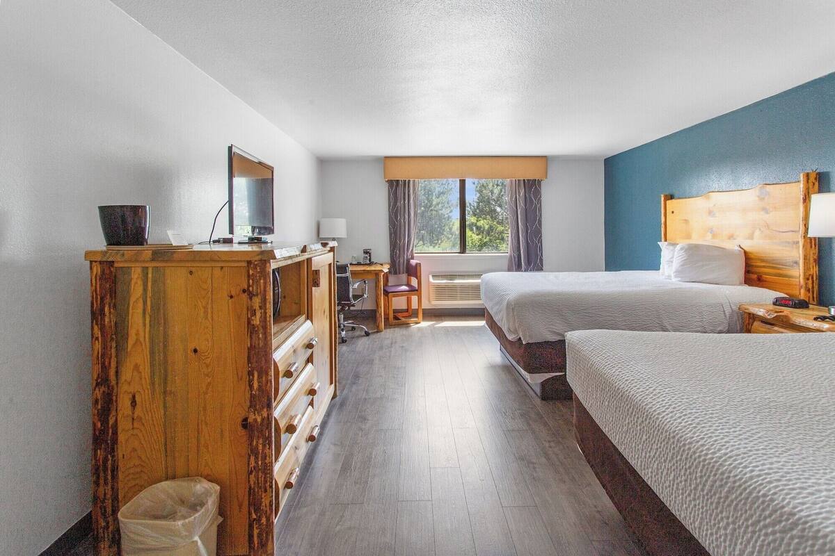 A hotel room with two beds, a wooden dresser, a desk with a chair, a window with curtains, and a view of trees.