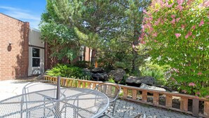 Une terrasse avec une table et des chaises en métal, entourée de verdure et bordée d’un bâtiment en briques.