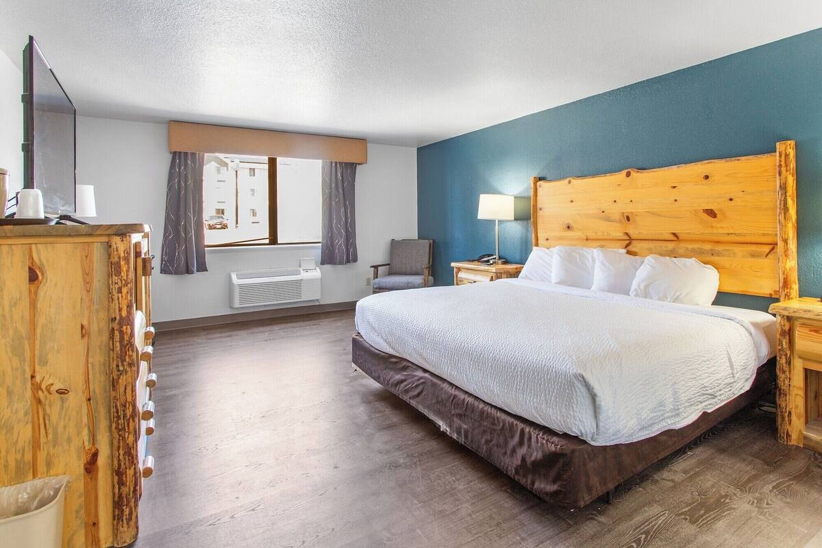 A hotel room with a wooden headboard, a large bed with white linens, a wooden dresser, a chair, and a window with curtains.