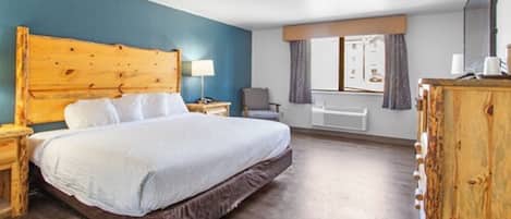 A hotel room with a large bed, wooden headboard, a desk with a lamp, a chair, and a window with curtains.