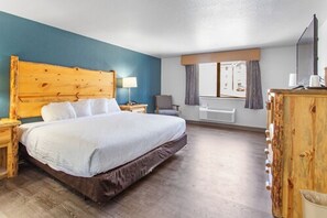 A hotel room with a large bed, wooden headboard, a desk with a lamp, a chair, and a window with curtains.