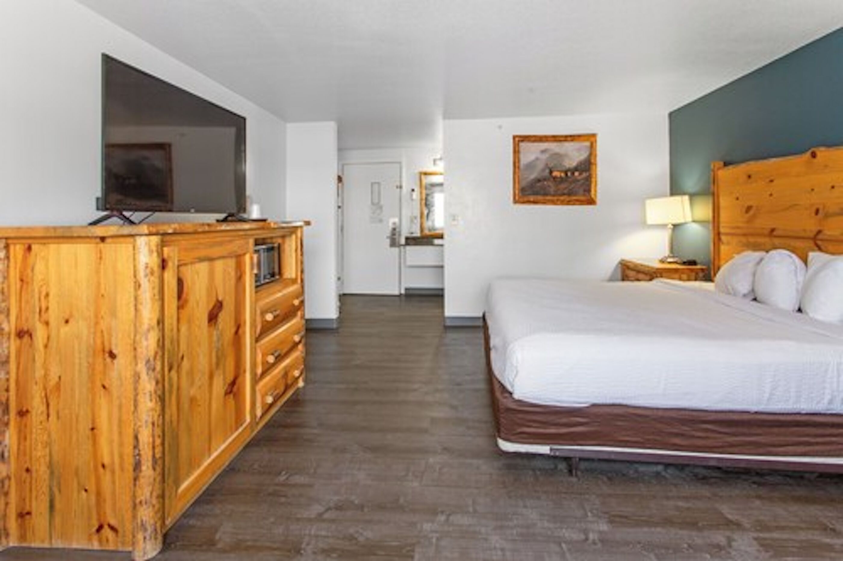 Une chambre avec une commode en bois, un lit avec une literie blanche, un téléviseur posé sur un meuble et un tableau accroché au mur.