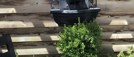 A black metal fountain with a fleur-de-lis design, surrounded by potted plants and a wooden fence.