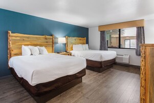 Habitación de hotel con dos camas, cabecero de madera, mesita de noche con lámpara, ventana con cortinas y equipo de aire acondicionado.