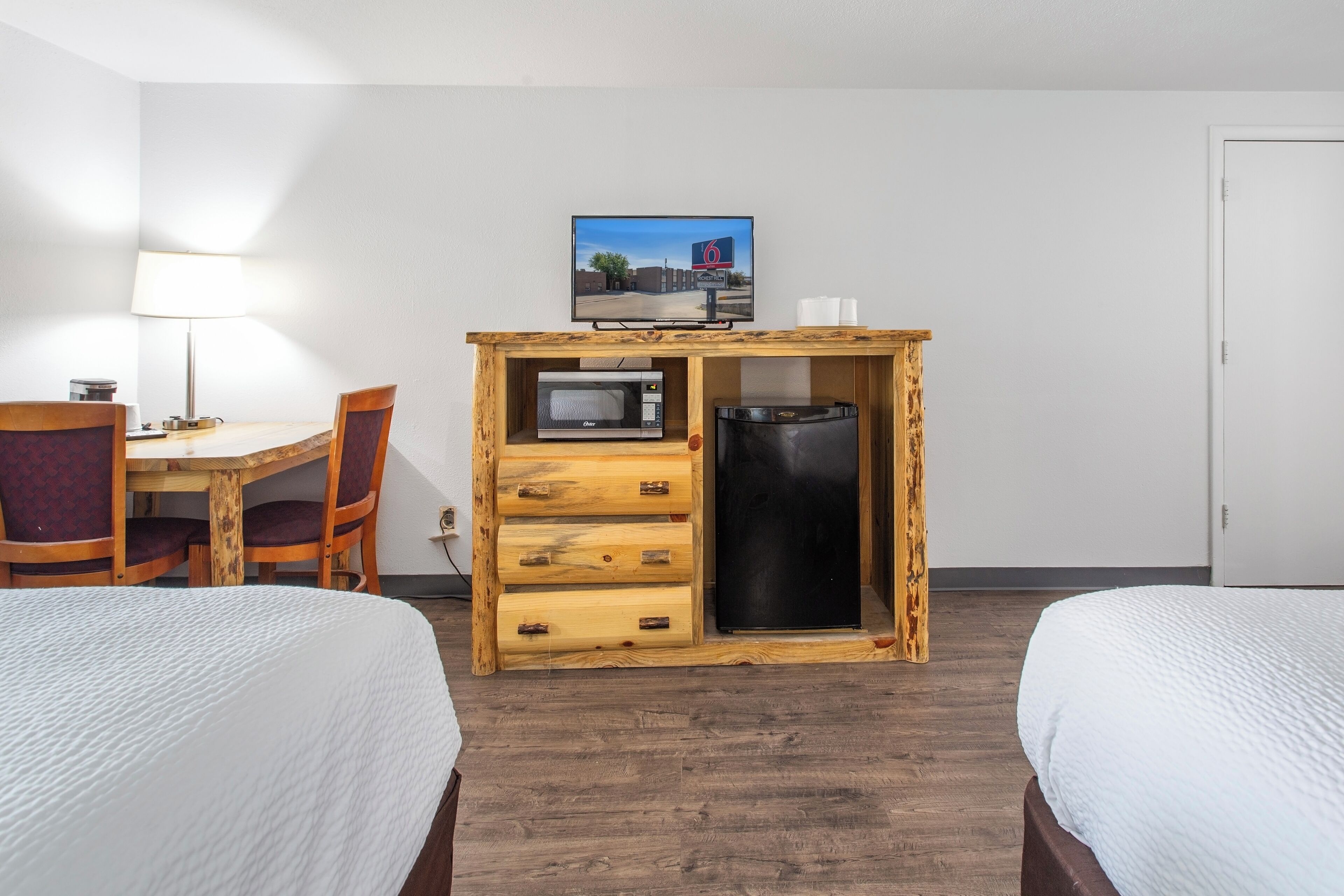 A hotel room with two beds, a wooden desk with a microwave, a refrigerator, a TV on the wall, and a lamp.