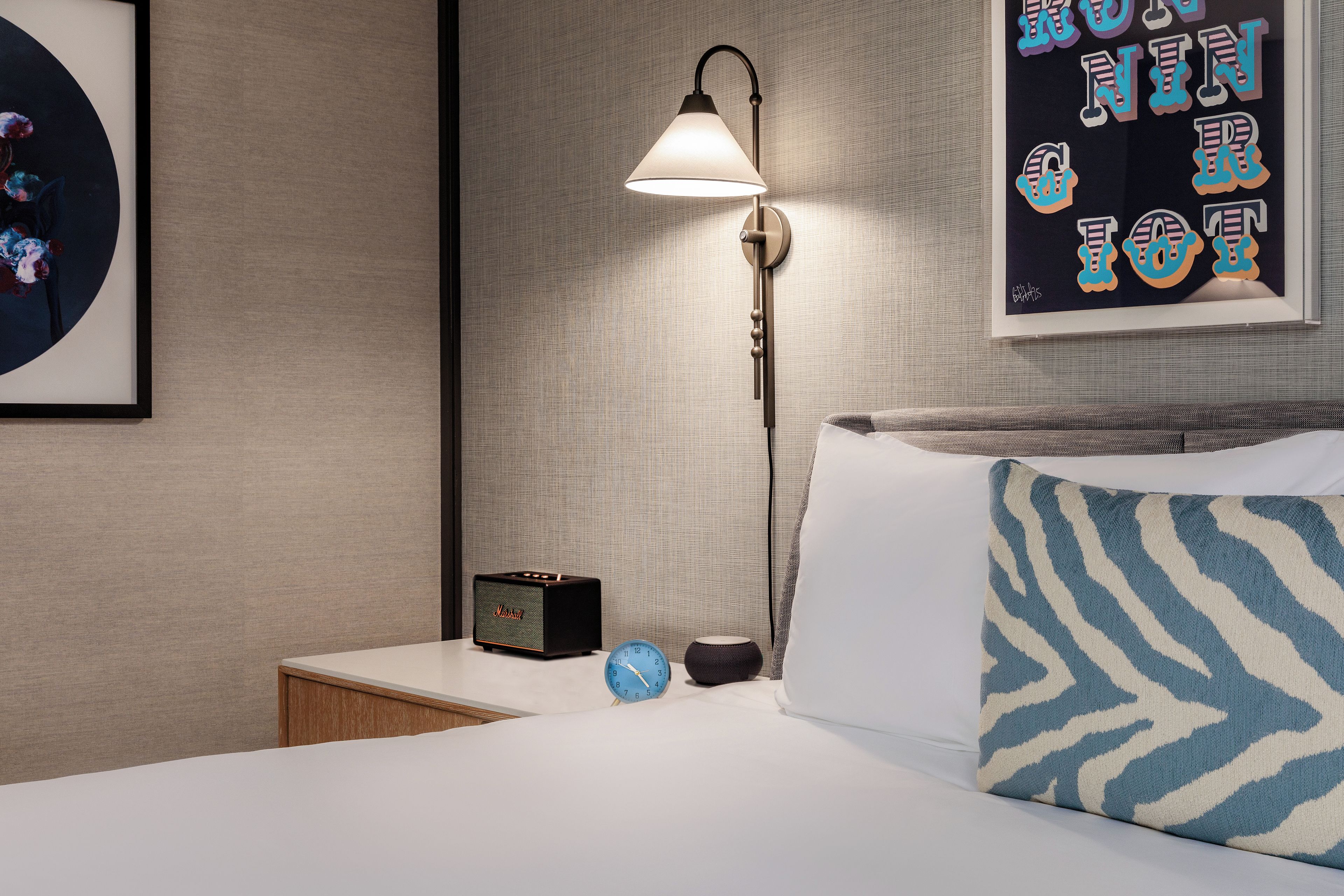 Habitación de hotel ordenada con una cama, una mesita de noche, una lámpara, un reloj y un cuadro en la pared.
