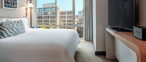 Ein Hotelzimmer mit einem großen Bett, einem Flachbildfernseher, einem Schreibtisch mit Lautsprecher und Blick auf Gebäude durch große Fenster.