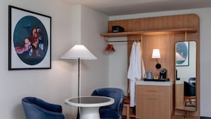 A hotel room with a round table, two blue chairs, a white lamp, a wooden cabinet, a mirror, and a framed abstract painting on the wall. - The Beacon at Embassy Row, WorldHotels Distinctive (Washington)