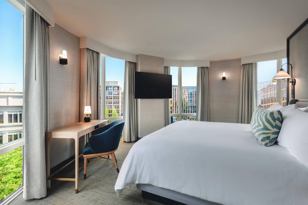 A hotel room with a bed, desk, chair, TV, and large windows with curtains.