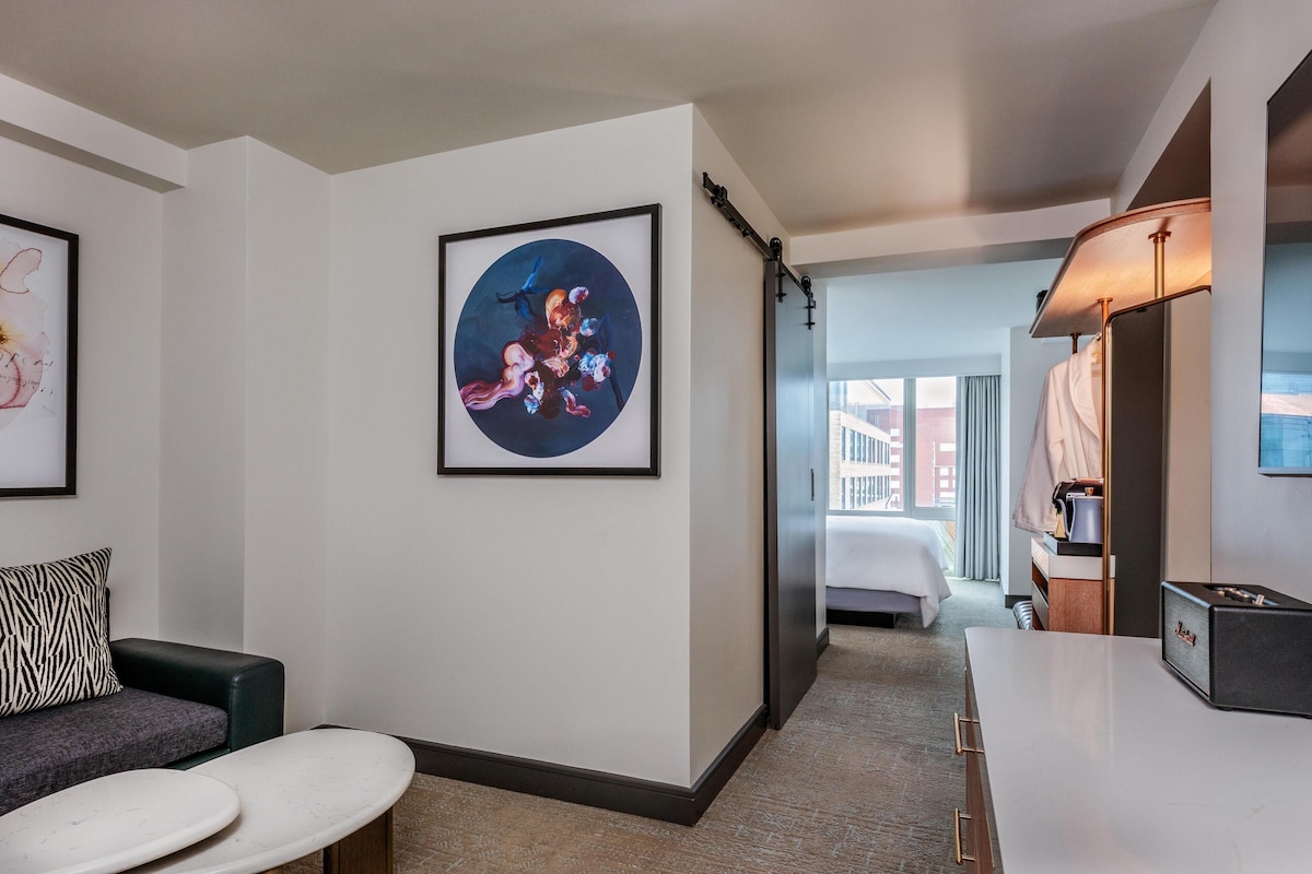 Habitación de hotel con cama, sofá, una mesita y un cuadro abstracto enmarcado en la pared.