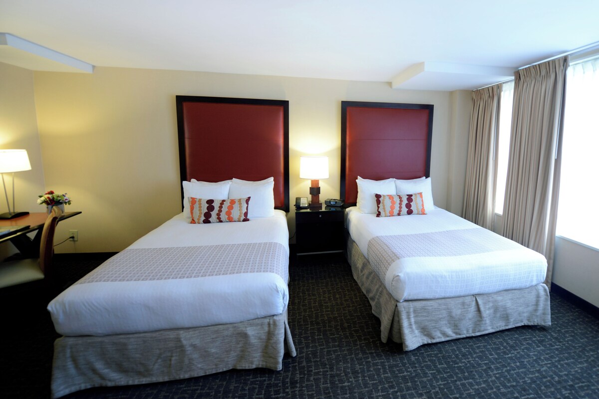 A hotel room with two beds, a desk, a lamp, and a window with curtains.