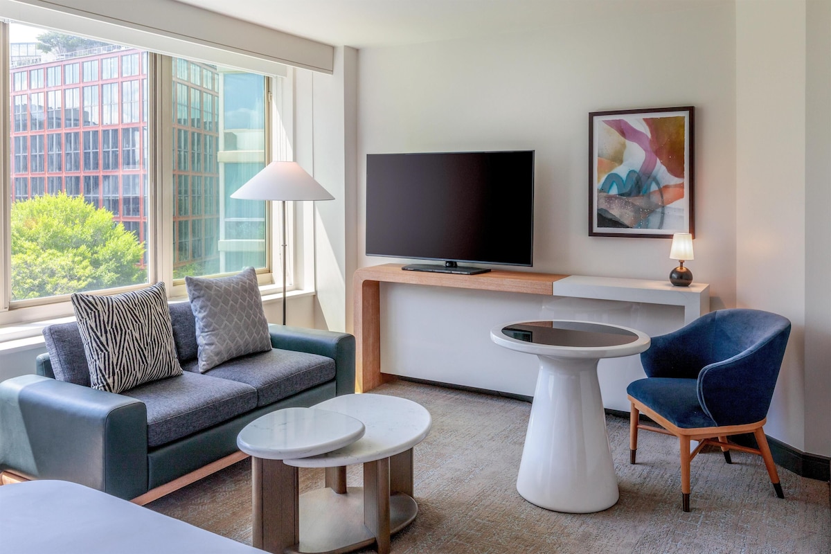 A modern hotel room with a sofa, a chair, a TV, a lamp, and a painting on the wall.