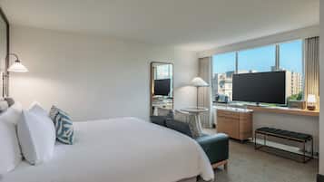 A hotel room with a bed, a desk, a chair, a bench, a TV, and a window with a city view.