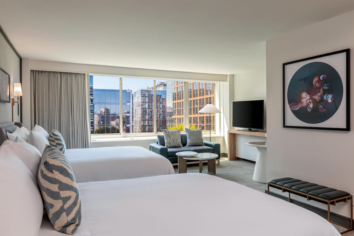 Habitación de hotel con dos camas, un sofá, un televisor, un cuadro y un ventanal con vistas a la ciudad.