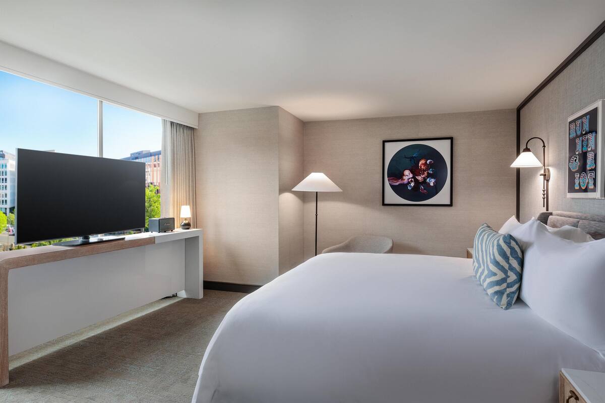 A hotel room with a large bed, a flat-screen TV on a stand, a desk with a lamp, and a city view.