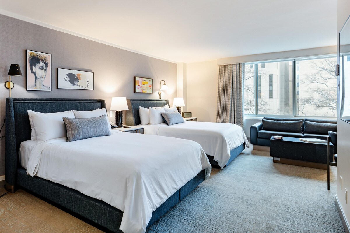 A hotel room with two beds, a sofa, a large window, and artwork on the walls.