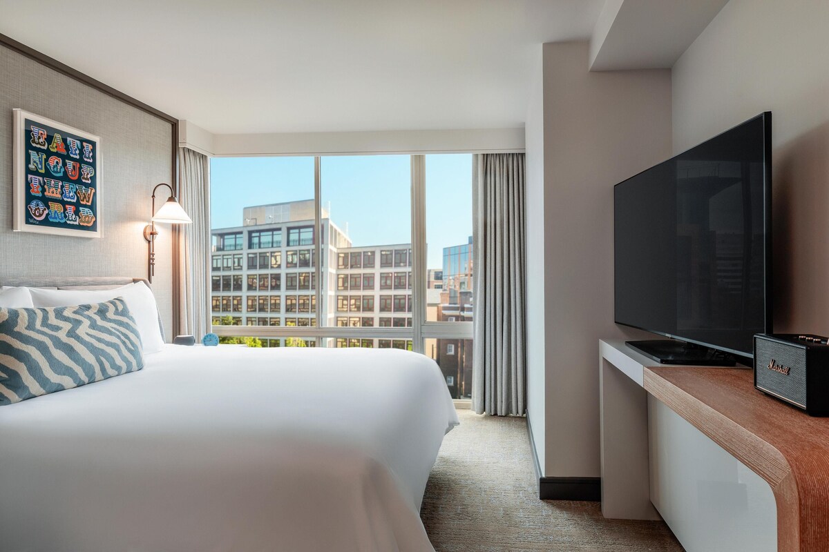 A hotel room with a large bed, a flat-screen TV, a desk with a speaker, and a view of buildings through large windows.