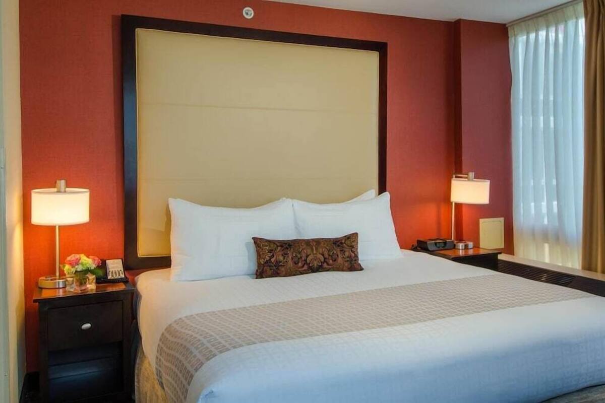 A hotel room with a large bed, two bedside tables with lamps, a nightstand, and a window with curtains.