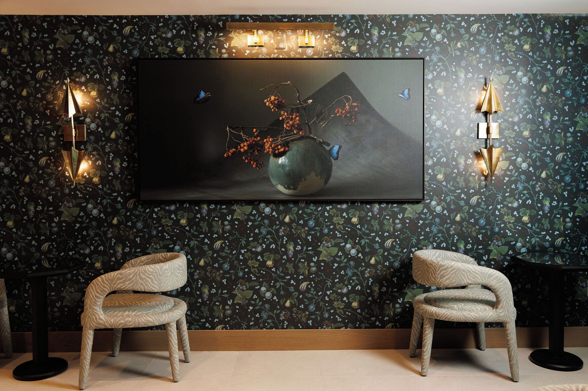 A room with a large painting of a vase with berries and butterflies, two chairs, and wall sconces.