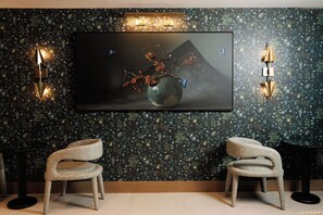 Une pièce décorée d’un grand tableau représentant un vase rempli de baies et entouré de papillons, ainsi que de deux chaises et d’appliques murales.